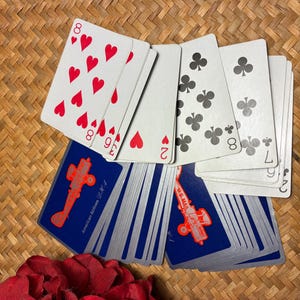 May include: A deck of playing cards with a blue backing featuring an orange airplane graphic and the text "American Airlines". Several cards are fanned out, showing hearts and clubs. The cards are on a woven surface.