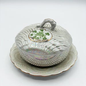 May include: A pearlescent, lidded dish with a matching saucer. The dish features a textured, ridged design and a small handle on top. A circular emblem with green shamrocks is on the lid. The saucer and dish have gold trim.