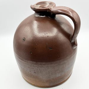 May include: A round, brown ceramic jug with a smooth, glossy finish. The vessel features a single curved handle attached to the side and a small, circular opening at the top with a slightly raised, molded rim. The surface shows subtle color variations.