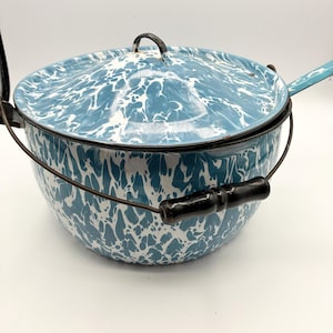 May include: A vintage blue and white enamelware pot with a lid. The pot has a speckled pattern and a black handle. The lid has a black handle as well. The pot has a wire handle and a long handle on the side. The pot is likely used for cooking.