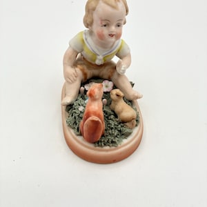 May include: A porcelain figurine of a seated child in a yellow shirt and brown shorts, with two small animal figures. The base is oval-shaped and features green foliage and small flowers. The figurine has a vintage aesthetic.