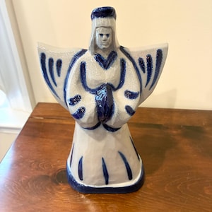 May include: A ceramic angel figurine with a light gray body and blue accents. The angel has outstretched wings and clasped hands, with blue detailing on the wings, robe, and base. The figurine is displayed on a wooden surface.