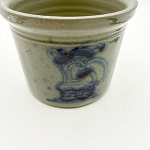 May include: A light gray ceramic crock with a speckled finish and a wide, flared rim. The exterior features a dark blue stamped graphic of a figure wearing a hat and scarf, positioned above a brick-patterned base, creating a textured appearance.