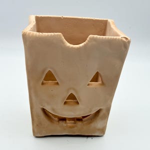 May include: A tan-colored, square-shaped Halloween decoration with a carved jack-o'-lantern face. The face features triangular eyes, a triangular nose, and a wide, toothy grin. The top edge has a curved cutout.