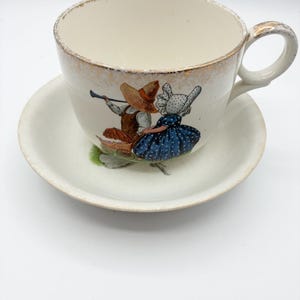 May include: Cream-colored teacup and saucer set with a gold rim. The cup features a painted scene of two figures in traditional attire. The saucer is round and matches the cup's color. A charming vintage tea set.