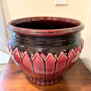 May include: A round ceramic planter featuring a glossy finish with a deep burgundy and black color palette. The exterior displays a raised, vertical petal-like pattern around the body, while the rim is smooth and wide with a dark, reflective glaze.