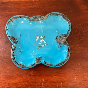May include: A turquoise-colored, four-lobed dish with a brown, textured rim. The dish has a glossy finish and features small floral accents in the center. The dish is likely made of ceramic or glass and is displayed on a wooden surface.