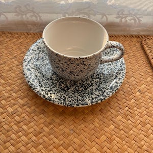 May include: A ceramic cup and saucer set featuring a speckled pattern of black dots on a white background. The cup has a rounded body with a curved handle, and the saucer is circular with a matching black and white speckled finish throughout.