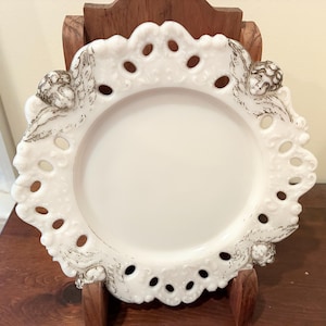 May include: A white, decorative plate with a scalloped edge and openwork design. The plate features small, embossed cherub figures and a raised rim. The plate is displayed on a wooden stand, showcasing its intricate details and vintage aesthetic.