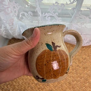 May include: A small, speckled ceramic pitcher with a beige base color. The pitcher features a hand-painted orange pumpkin design on the front, complete with a green leaf and brown stem. It has a curved handle and a flared rim with a small pouring spout.