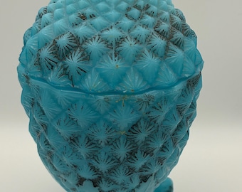 May include: A vintage, light blue, pineapple-shaped trinket box. The glass box has a textured surface with a detailed pattern and rests on three small feet. The lid has a ruffled edge.