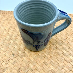 May include: A light blue ceramic mug with a cylindrical shape and horizontal ribbed texture on the interior. The exterior features a dark blue hand-painted grape vine motif with clusters of dots representing grapes and curved stems.