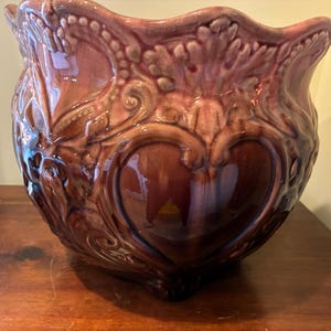 May include: A large, decorative ceramic planter with a glossy finish. The planter is a deep burgundy color with embossed floral and heart-shaped designs. The rim has a scalloped edge.