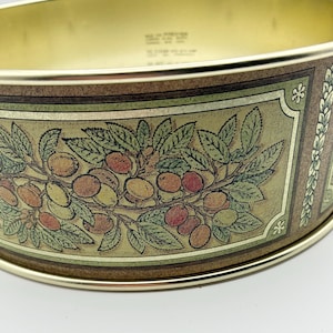 May include: A vintage metal planter with a gold rim and a decorative band featuring a botanical design. The band showcases a branch with green leaves and various colored fruits, including red, orange, and yellow. The planter has a curved shape and a handle.