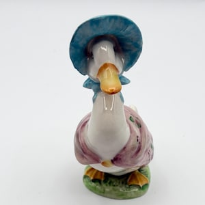 May include: A ceramic figurine of a goose wearing a blue bonnet and a pink and purple dress. The goose has a yellow beak and orange feet, standing on a green base. A charming decorative item.