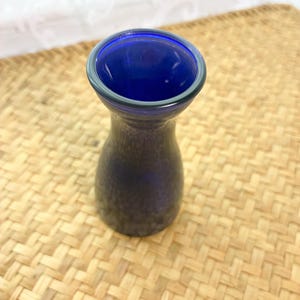 May include: A small, cobalt blue glass vase with a flared rim and a smooth, rounded body. The glass features a deep, saturated blue hue that is consistent throughout the piece, with a clear, circular opening at the top and a tapered neck.