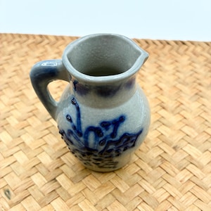 May include: A small, light gray ceramic pitcher with a single handle and a pouring spout. The exterior features a textured, cobalt blue floral design embossed on the front, with additional blue glaze accents around the rim and handle base.