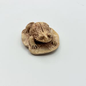 May include: A small, beige, textured figurine of a frog resting on a lily pad. The frog has a bumpy, detailed surface with visible eyes and limbs, while the lily pad base features etched lines representing veins, creating a realistic appearance.
