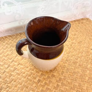 May include: A ceramic pitcher featuring a two-tone design with a dark brown glazed upper section and a cream-colored lower body. The pitcher includes a curved handle on the side and a distinct pouring spout at the rim, showcasing a smooth finish.