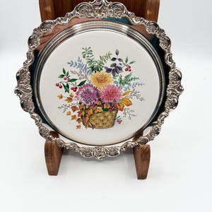 May include: A silver-plated serving tray with a floral design. The tray features a white center with a colorful illustration of flowers in a basket. The tray has an ornate, scalloped edge and is displayed on a wooden stand.
