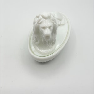 May include: A white ceramic trinket box featuring a sculpted lion head on the lid. The container is oval-shaped with a smooth, glossy finish. The lion is depicted in a resting position with its mane detailed and its head facing forward.