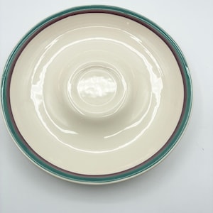 May include: A round, off-white ceramic serving dish featuring a raised center section. The rim is decorated with two concentric bands, one in a deep teal green and the other in a muted plum purple, creating a simple, clean border design.
