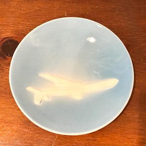 May include: A light blue ceramic plate with a raised, cream-colored airplane design. The plate has a circular shape and is set against a dark wood surface. The airplane is in flight.
