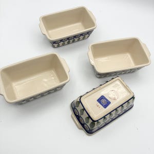 May include: Three rectangular ceramic loaf pans with cream-colored interiors and exterior patterns featuring blue circles and green leaf-like shapes. One pan is inverted to show the base, which includes a blue rectangular sticker and a circular stamp.