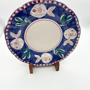 May include: A round ceramic plate with a scalloped edge featuring a central cream-colored circle surrounded by a deep blue border. The border is decorated with four hand-painted white fish with red details and green seaweed accents, finished with a red and white dotted rim.
