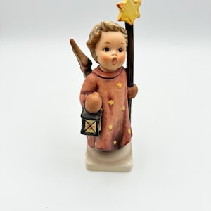 May include: A ceramic angel figurine with a surprised expression, wearing a reddish-brown robe adorned with yellow stars. The angel holds a black lantern in one hand and a star-topped staff in the other. The figurine is on a cream-colored base.
