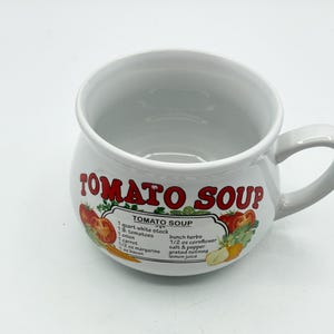 May include: A white ceramic mug with a rounded body and a handle. The front features the red text TOMATO SOUP above a recipe label listing ingredients including 1 quart white stock, 1 lb. tomatoes, 1 onion, 1 carrot, 1/2 oz margarine, 1 oz bacon, herbs, 1/2 oz cornflour, salt, pepper, nutmeg, and lemon juice.