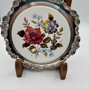 May include: A decorative serving tray with a floral design. The tray features a white center with a colorful flower arrangement, including a large red rose. It has a silver-colored, ornate rim and a teal-colored border. The tray is supported by a wooden stand.