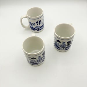 May include: Three white ceramic mugs featuring a blue willow pattern. Each mug has a cylindrical shape with a handle on the side. The blue design depicts traditional landscape scenes with pagodas, trees, and bridges encircling the exterior surface.