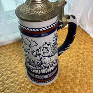 May include: A ceramic beer stein featuring a metal lid with an ornate embossed pattern. The body is white with blue and red accents, displaying a raised relief of a moose in a forest setting. The handle is dark blue with a twisted rope design.