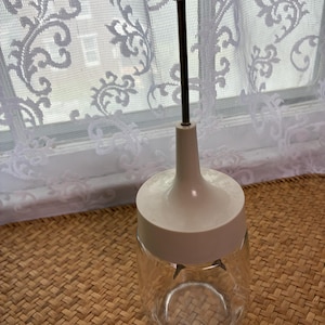 May include: A clear glass jar with a white plastic lid and a central plunger mechanism. The plunger features a thin metal rod extending upward to a flat white circular handle. The lid assembly includes a white conical base that fits onto the jar.