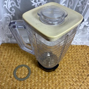 May include: A clear plastic blender pitcher with a square base and a beige lid featuring a removable clear center cap. The pitcher has a side handle and measurement markings in cups. A separate grey circular gasket ring sits on the surface nearby.