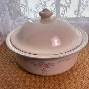 May include: A round, cream-colored ceramic casserole dish with a matching domed lid featuring a central knob. The exterior of the bowl is decorated with a delicate floral pattern consisting of soft pink roses and muted green leaves encircling the base.