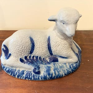 May include: A ceramic lamb figurine in a reclining pose. The lamb is primarily white with blue accents, including decorative markings on its body and a base depicting grass. The figurine is a decorative item.