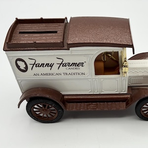 May include: A vintage-style toy truck in brown, white, and gold. The truck features the "Fanny Farmer Candies" logo and the text "An American Tradition" on its side. The truck has a slot on top, suggesting it may be a bank.