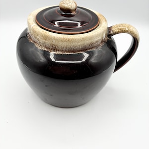 May include: A vintage ceramic crock with a lid. The crock is a glossy dark brown with a tan and brown rim and handle. The lid has a small knob. The crock is on a white background.