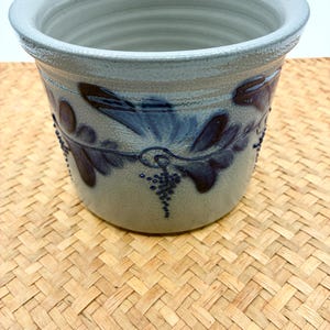 May include: A cylindrical ceramic crock with a light gray speckled glaze and a wide, flared rim. The exterior features a dark blue embossed floral pattern with leaf motifs and small dotted clusters encircling the body of the vessel.