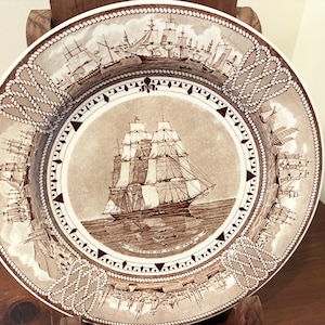 May include: A round ceramic plate featuring a sepia-toned illustration of a large sailing ship in the center with the text Witch of the Wave. The border is decorated with a repeating pattern of nautical ropes and smaller sailing vessels.