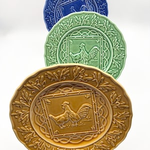 May include: A stack of decorative ceramic plates in blue, green, and gold. Each plate features a rooster and hen design within a square frame, surrounded by a leafy border. The plates have scalloped edges and are displayed on a metal stand.