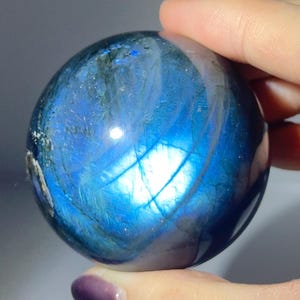 May include: A spherical labradorite gemstone, predominantly blue with iridescent flashes of teal and gold. The polished orb is held in a hand, highlighting its reflective surface. This gemstone is a popular choice for collectors and those interested in crystals. The stone's diameter is approximately 5 cm.