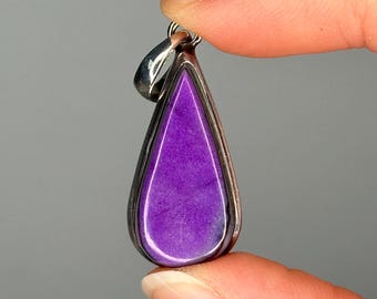 May include: A teardrop-shaped pendant with a vibrant purple stone set in a silver-colored metal frame. The pendant is held between two fingers against a gray background. The stone has a textured appearance.