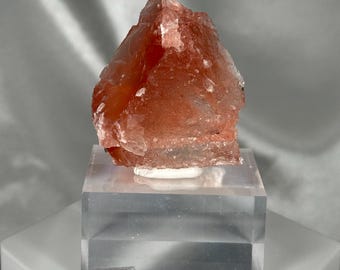 May include: A rough, reddish-orange crystal formation, possibly a mineral specimen, is displayed on a clear acrylic block. The crystal has a textured surface and is mounted on a small white base. The background is a soft, neutral gray.