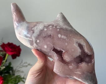 May include: A pink and white crystal dolphin sculpture. The sculpture has a smooth, polished exterior with a geode-like interior. The dolphin's form is streamlined, with a pointed dorsal fin and a curved body. The crystal has a natural, organic appearance.