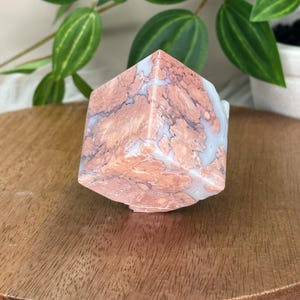 May include: A geometric crystal with a mix of peach and white tones. The stone has a unique pattern and is displayed on a wooden surface. Green leaves and a white pot are in the background.