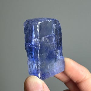 May include: A striking, translucent blue crystal with a rectangular form. The crystal has a rough, textured surface and is set against a grey backdrop. The crystal's colour varies from deep indigo to lighter shades of blue.
