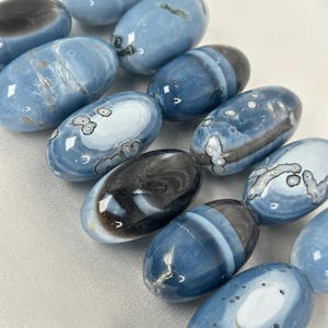 May include: A selection of oval blue and black gemstones. The stones display varying shades, with some featuring bands of black and white. The polished stones are arranged on a white surface, highlighting their natural patterns and textures.